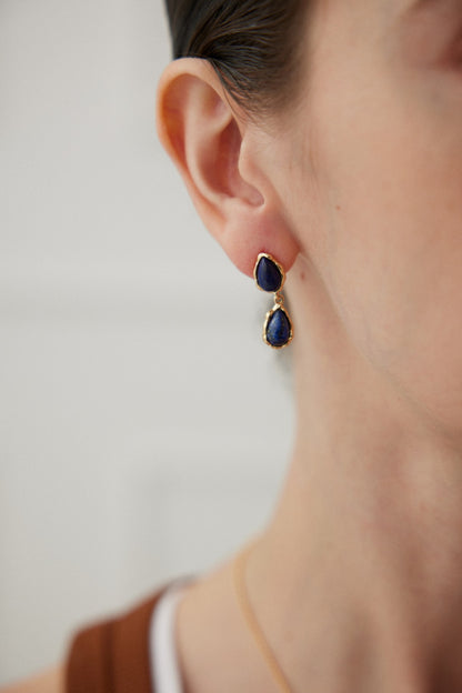 Close-up of an ear adorned with Siren Mode's Elegant Lapis Lazuli Teardrop Earrings in Vintage Gold. Each earring features two dark blue stones set in a vintage gold setting. The softly blurred background highlights the exquisite jewelry.