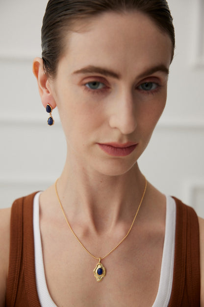 A person with fair skin, brown hair, and light blue eyes wears Siren Mode's Elegant Lapis Lazuli Teardrop Earrings in Vintage Gold and a matching gold necklace with a blue gemstone pendant. They are wearing a brown shirt with a white tank top underneath.