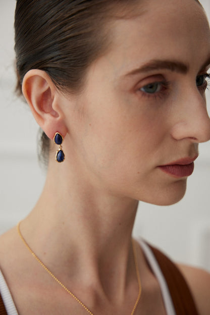 Close-up of a person wearing Siren Mode's Elegant Lapis Lazuli Teardrop Earrings in Vintage Gold and a delicate gold necklace. The individual has light skin, dark slicked-back hair, and is seen in profile against a blurred, neutral background.