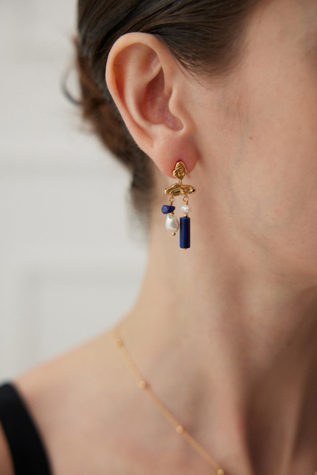 Close-up of a person wearing the Siren Mode "Elegant Lapis Lazuli and Pearl Drop Earrings in Vintage Gold." The gold accessory is accentuated with blue, white, and coral-colored beads. The person has short hair, and the background is blurred, highlighting the earring.