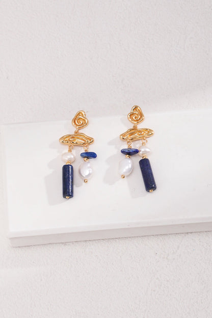 The Elegant Lapis Lazuli and Pearl Drop Earrings in Vintage Gold by Siren Mode feature a unique twisted design at the top, with rectangular lapis lazuli stones and white pearls. These exquisite jewelry pieces are elegantly displayed on a white surface against a light-colored background.