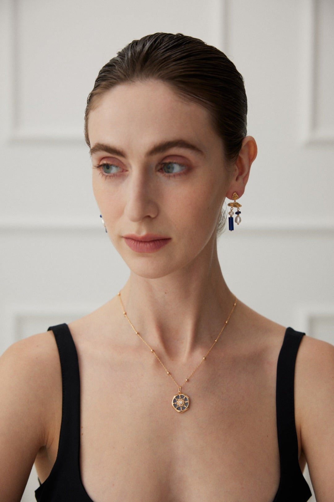 A woman with light skin and slicked-back hair is wearing a black sleeveless top. She is accessorized with a gold necklace featuring a circular pendant and the stunning Elegant Lapis Lazuli and Pearl Drop Earrings in Vintage Gold by Siren Mode. The background is a softly lit white wall with panel details.