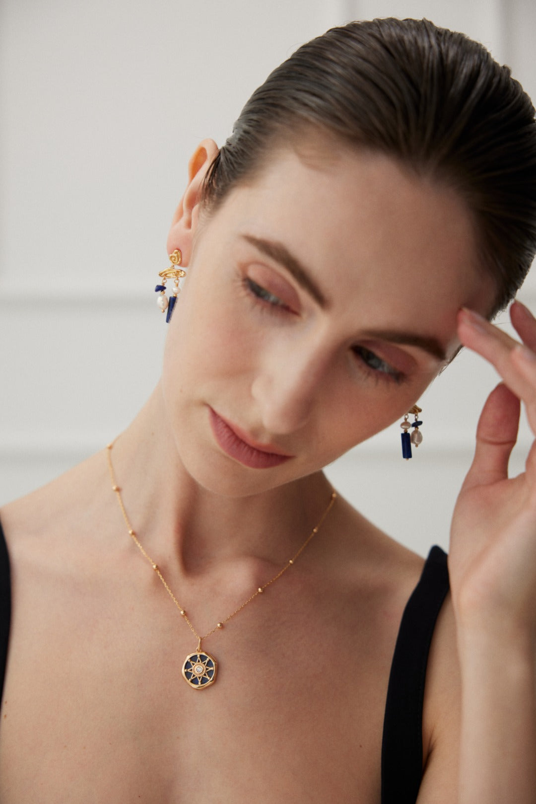 A woman with slicked-back hair wearing a black top, a gold pendant necklace with a circular design, and matching Elegant Lapis Lazuli and Pearl Drop Earrings in Vintage Gold by Siren Mode. She is looking down, touching the side of her head with one hand against a blurred, light-colored background.