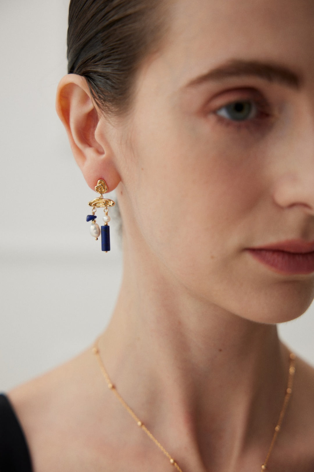 A close-up of a person wearing Siren Mode's Elegant Lapis Lazuli and Pearl Drop Earrings in Vintage Gold. The earrings feature both a pearl and a blue rectangular lapis lazuli charm. The person's face is partially visible, with sleek, pulled-back hair and a hint of a green eye shown. They also adorn a dainty gold necklace.