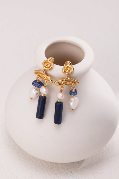 A pair of Elegant Lapis Lazuli and Pearl Drop Earrings in Vintage Gold by Siren Mode is elegantly displayed on a smooth, white ceramic vase. These earrings feature blue lapis lazuli beads, freshwater pearls, and a unique spiral gold wire design at the top. The contrast between the colorful beads and the gold wirework is striking in this exquisite jewelry piece.