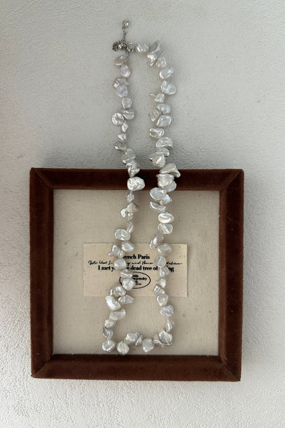 The Elegant Natural Keshi Pearl Necklace in Sterling Silver from Siren Mode is beautifully showcased against a backdrop of fabric within a square frame bordered by brown velvet. The fabric features partially legible text that reads "Paris" and "dead tree," which accentuates the organic charm of the necklace.