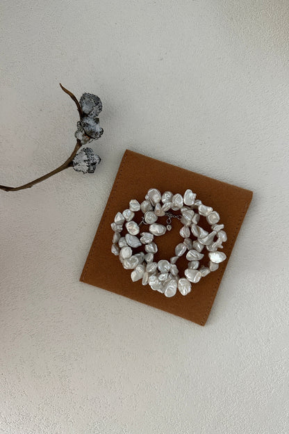 The Elegant Natural Keshi Pearl Necklace in Sterling Silver by Siren Mode is beautifully showcased on a brown square card. Accompanying the necklace is a slender branch adorned with three dried, dark-hued seed pods or berries, all set against a white textured background that accentuates its organic charm.