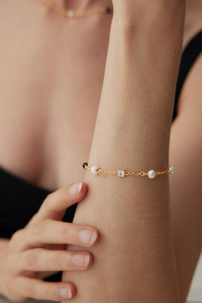 A person wearing the Elegant Pearl, Onyx, and Zirconia Bracelet - Vintage Gold by Siren Mode. The bracelet is adorned with small pearls and a central black onyx gemstone. The person is dressed in a black top, with only the lower part of their face and upper torso visible, while one hand gently touches the bracelet on the other arm.