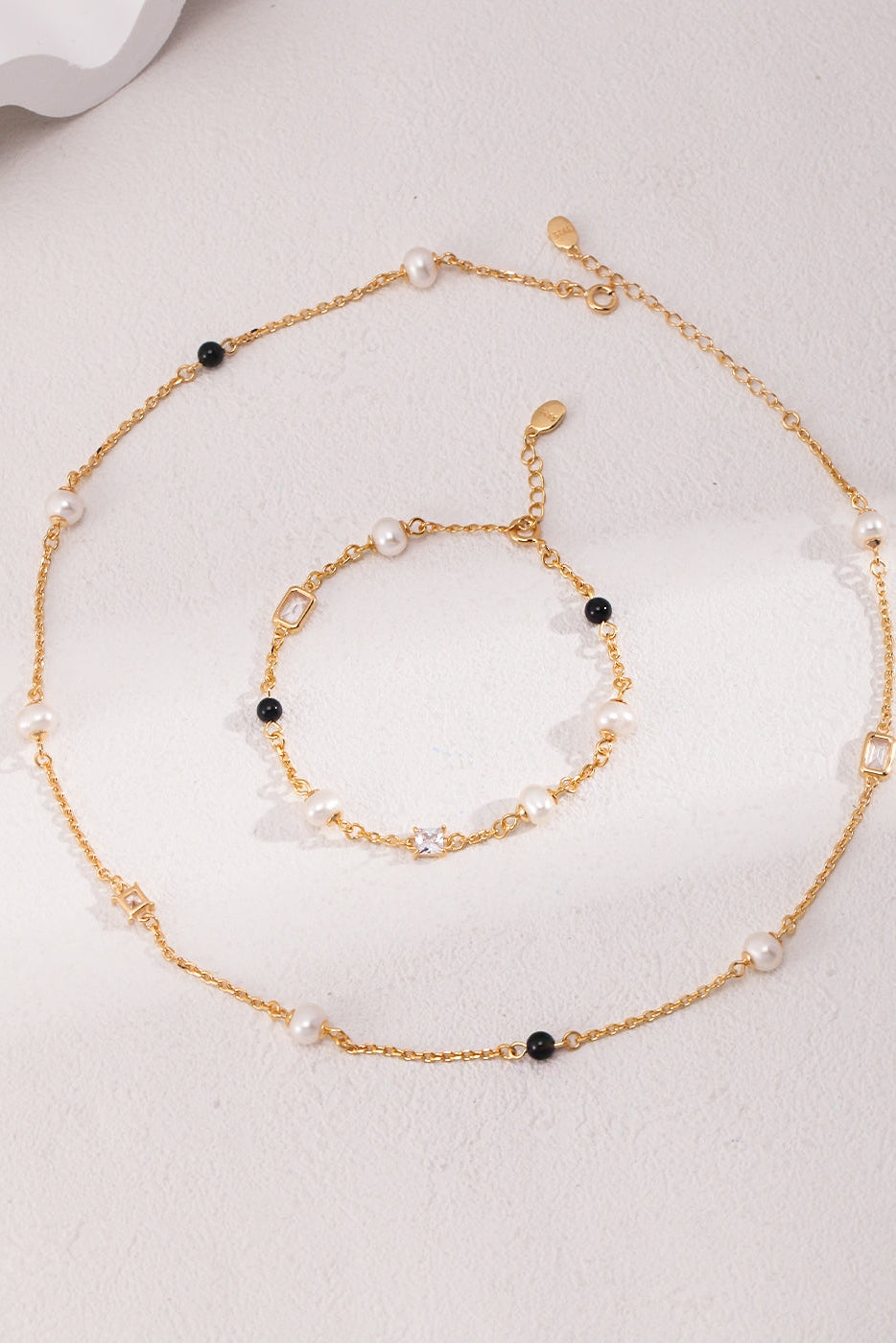 A matching set of delicate gold chain jewelry consisting of a necklace and the Elegant Pearl, Onyx, and Zirconia Bracelet - Vintage Gold by Siren Mode, each adorned with small white pearls, black onyx, and beads. The jewelry is arranged neatly on a smooth, light-colored surface with the necklace lying in a circular shape alongside the bracelet.