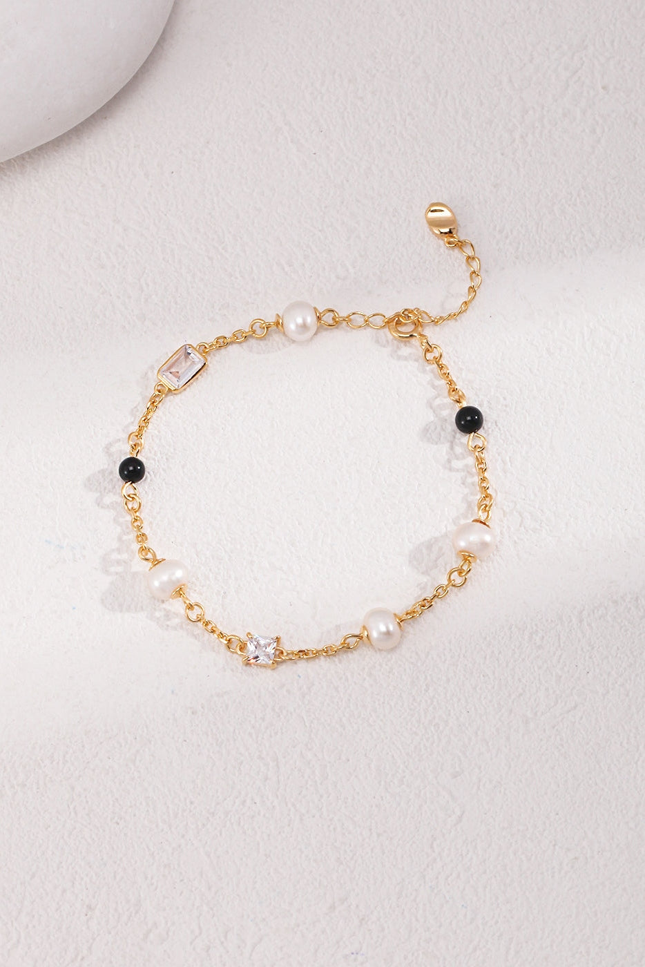 The Siren Mode Elegant Pearl, Onyx, and Zirconia Bracelet - Vintage Gold is a delicate bracelet featuring white pearls, black onyx beads, a small square charm, and a star-shaped zirconia gem charm arranged in a linear pattern. It has an adjustable chain with a lobster clasp and includes a tiny flat charm at the end. The bracelet is displayed on a light surface.