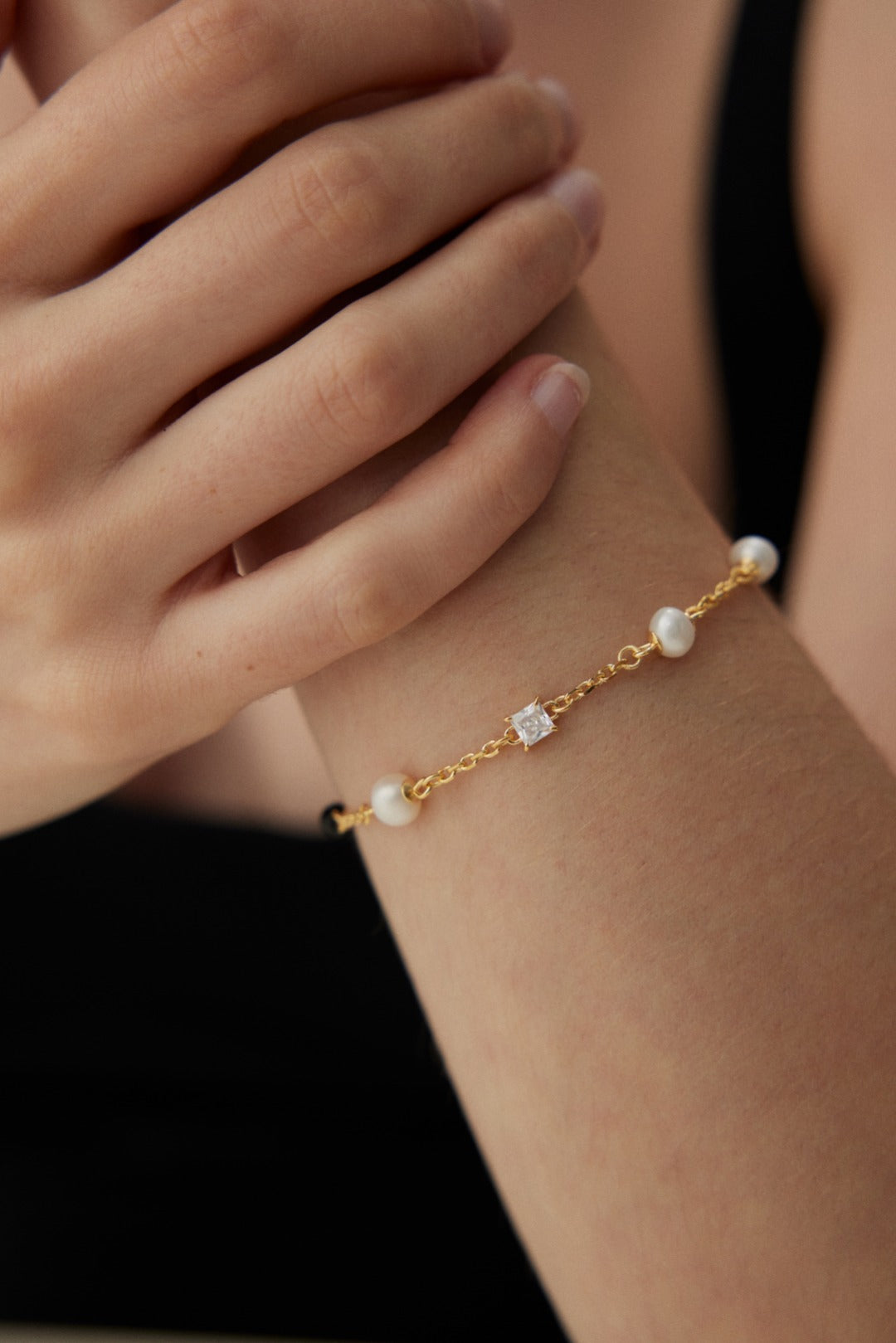 Close-up of a person wearing the Siren Mode Elegant Pearl, Onyx, and Zirconia Bracelet - Vintage Gold. The delicate bracelet is adorned with small white pearls and a square gemstone. The person's hand gently rests on their forearm, showcasing the bracelet. They are dressed in a black top.