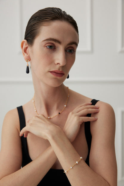 A woman with light skin and dark hair pulled back is wearing black drop earrings, a delicate necklace, and the Siren Mode Elegant Pearl, Onyx, and Zirconia Bracelet - Vintage Gold. She is dressed in a black sleeveless top and is posed with hands gracefully held near her chest.