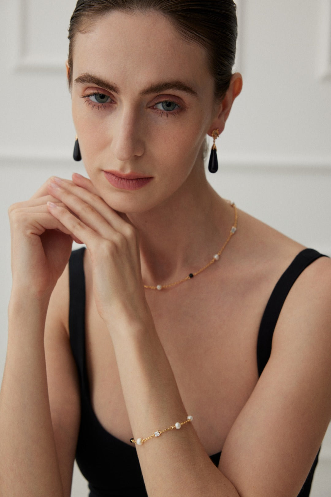 A woman with fair skin and slicked-back hair is wearing a black sleeveless top. She is adorned with matching zirconia jewelry, including black drop earrings, a delicate necklace, and the Elegant Pearl, Onyx, and Zirconia Bracelet - Vintage Gold by Siren Mode. She rests her chin gently on her clasped hands, looking contemplative.