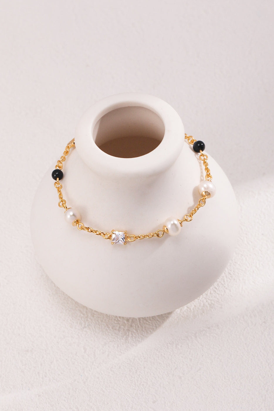 An Elegant Pearl, Onyx, and Zirconia Bracelet - Vintage Gold by Siren Mode drapes elegantly around the narrow neck of a white, minimalist vase against a textured light background, showcasing its delicate gold design adorned with small white pearls, black onyx beads, and a central white gemstone.