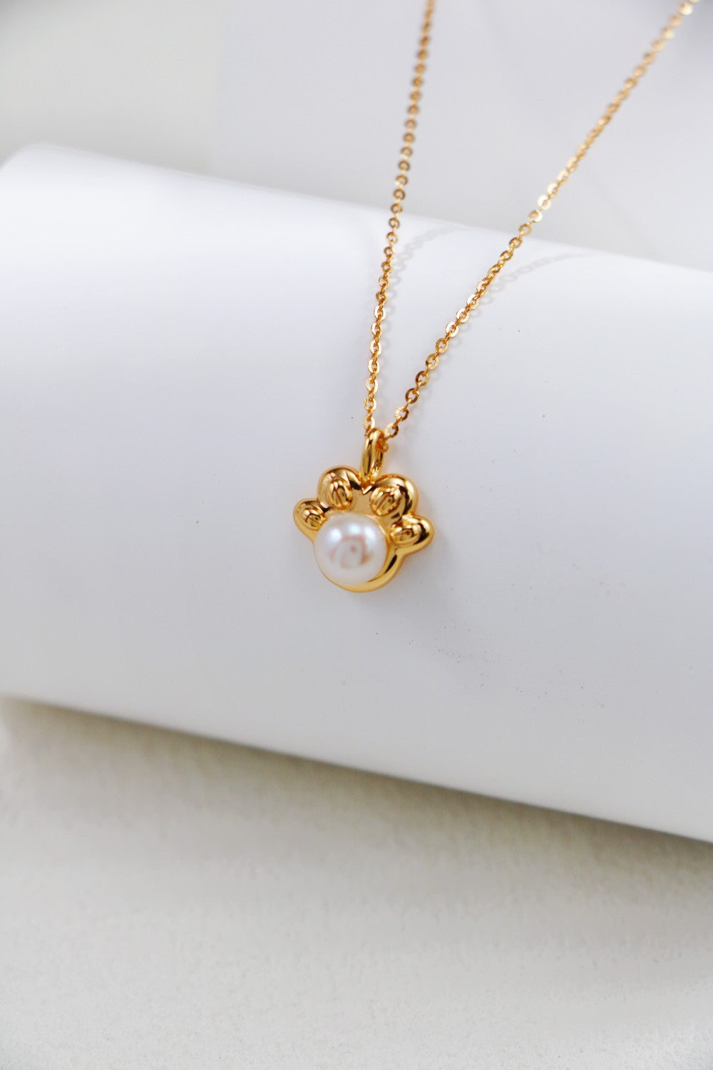 The Elegant Pearl Pendant Necklace by Siren Mode, crafted from sterling silver and adorned with a charm resembling a paw print, features a delicate white pearl at its center and is draped over a white cylindrical object. The softly blurred background beautifully highlights the pearl pendant.