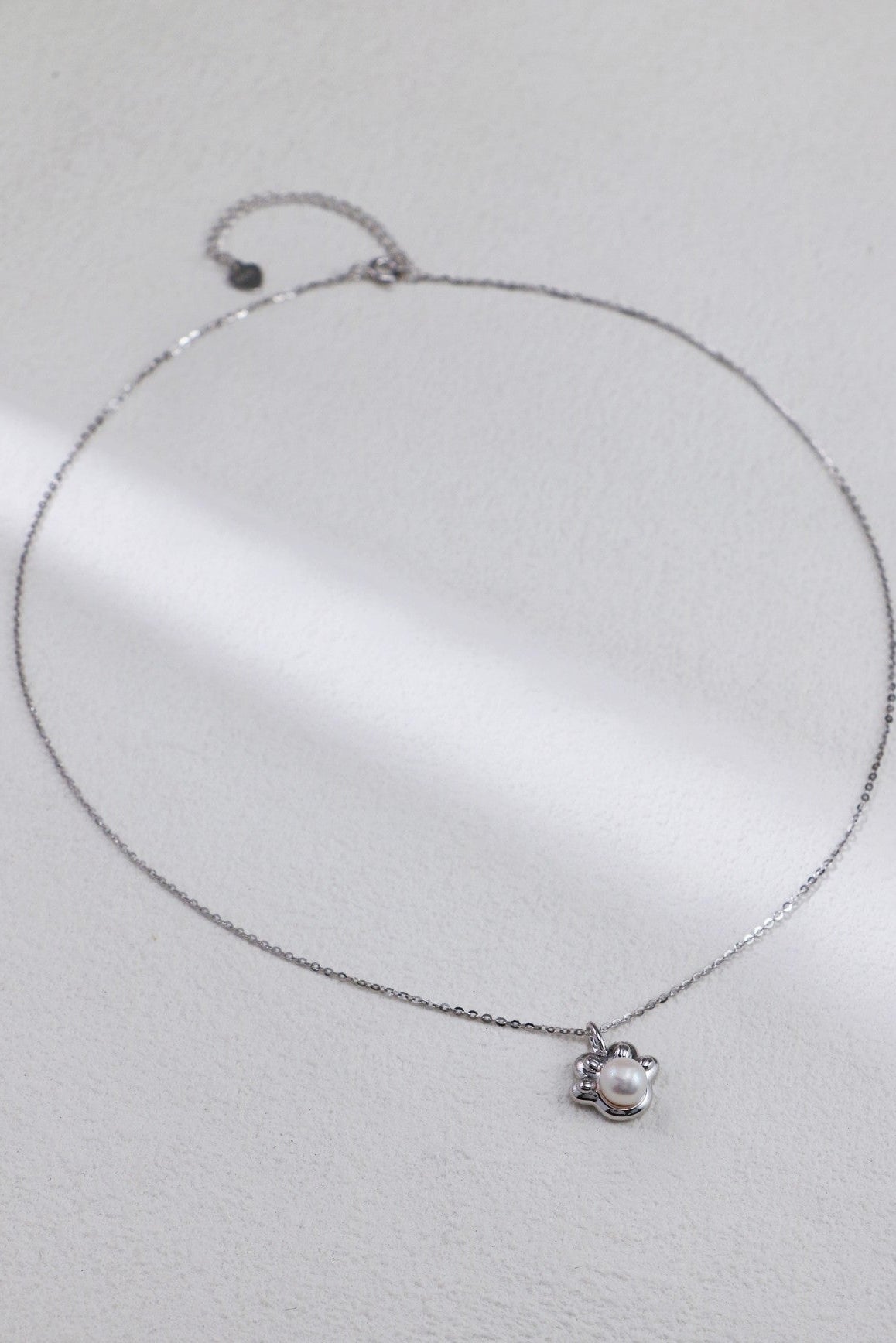 An Elegant Pearl Pendant Necklace in Sterling Silver by Siren Mode, featuring a delicate chain and a small pearl pendant. The pendant showcases a single, round pearl encased in an ornate silver setting and is photographed against a plain, light-colored background.