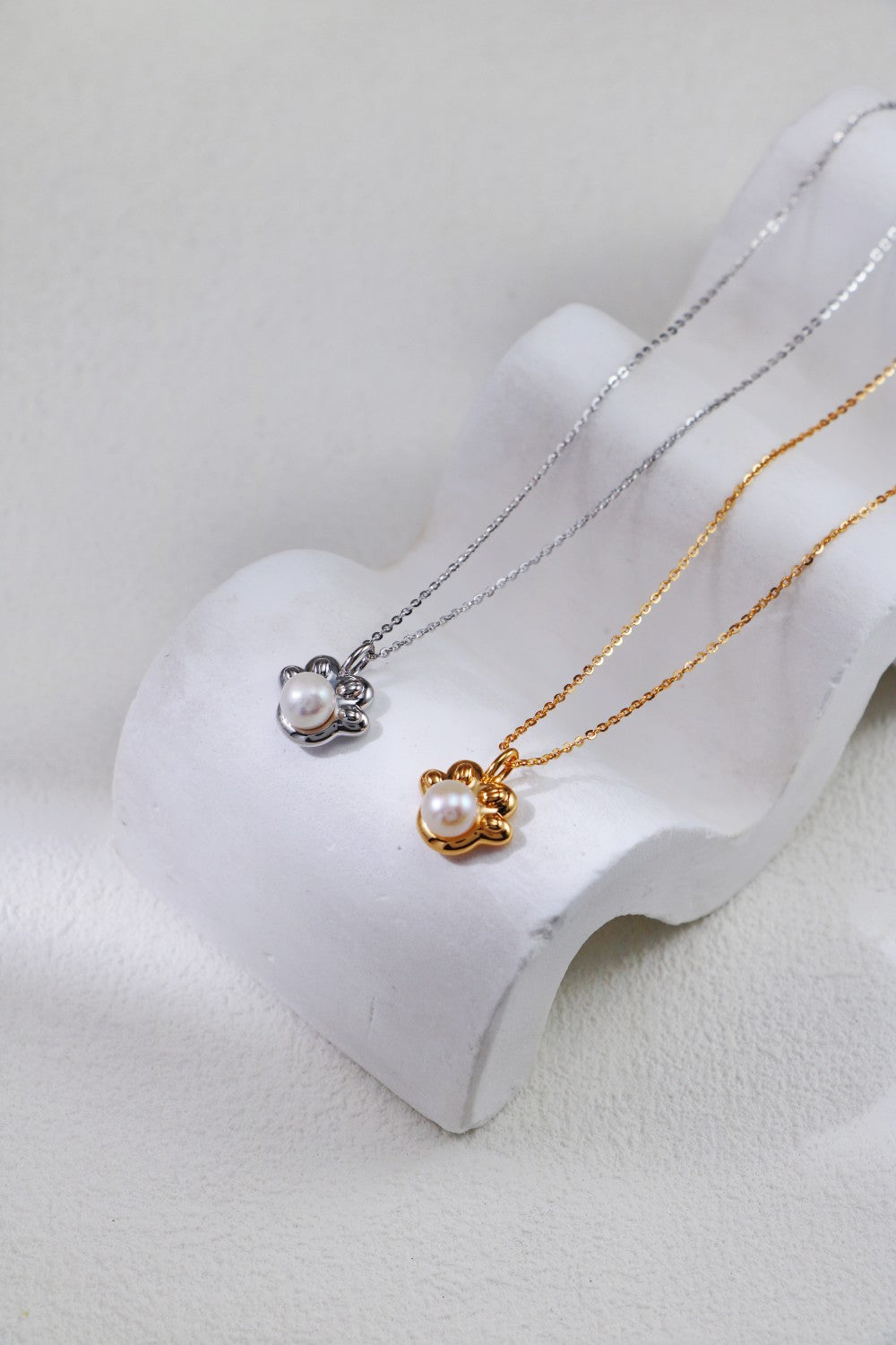 Two necklaces rest on a white, wave-like display stand. Each features a paw print pearl pendant at its center. One is an Elegant Pearl Pendant Necklace in vintage gold, while the other is the Siren Mode Elegant Pearl Pendant Necklace in Sterling silver. The neutral background emphasizes the elegance of the jewelry.