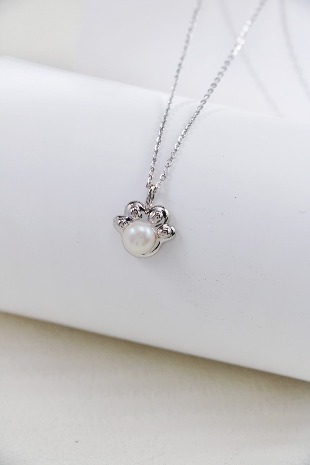 The Elegant Pearl Pendant Necklace - Sterling Silver by Siren Mode is a delicate piece featuring a paw print-shaped pendant. The paw print includes a central pearl and is accented with small, intricate detailing. The necklace is beautifully displayed on a white, cylindrical backdrop.