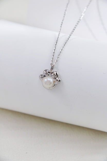 The Elegant Pearl Pendant Necklace - Sterling Silver by Siren Mode is a delicate piece featuring a paw print-shaped pendant. The paw print includes a central pearl and is accented with small, intricate detailing. The necklace is beautifully displayed on a white, cylindrical backdrop.