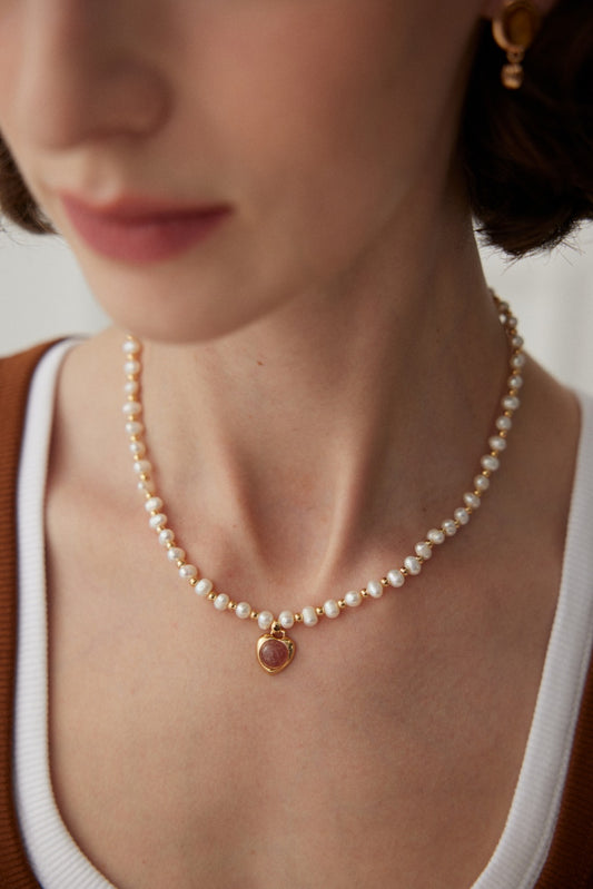 A close-up shot of a person wearing Siren Mode's Elegant Pearl and Strawberry Quartz Pendant Necklace in Vintage Gold with a white and brown top. The focus is on the natural pearls, showcasing their details alongside the elegance of the 925 silver necklace.