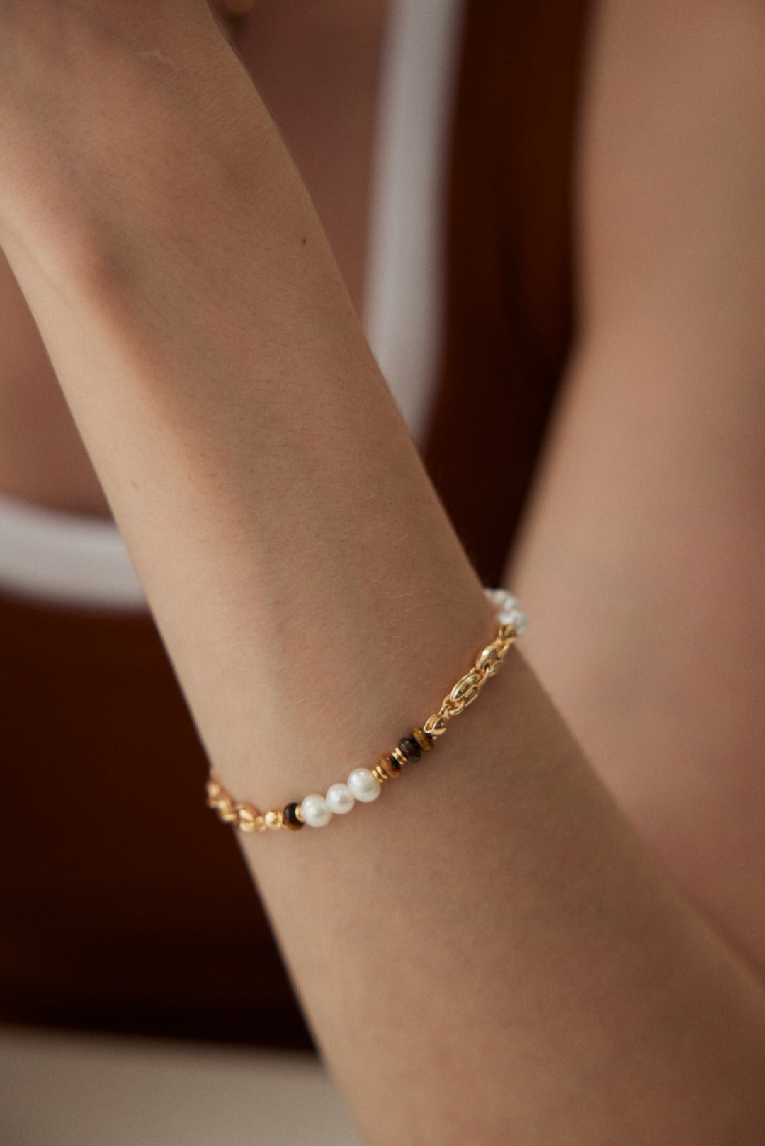 Close-up of a person's arm wearing the Elegant Tiger's Eye and Pearl Bracelet in Vintage Gold by Siren Mode. The bracelet, featuring natural pearls and vintage gold links, adds an elegant touch to the minimalist and stylish appearance. The person is wearing a sleeveless top.