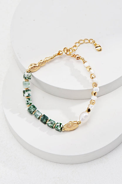 The Elegant Vintage Gold Bracelet by Siren Mode features green marbled Qinghai jade rectangular beads, gold beads, and a single white bead, all elegantly complemented by a gold chain and clasp. Displayed on a white, tiered circular platform, this bracelet also incorporates pearl and mother of pearl accents set in 925 silver.