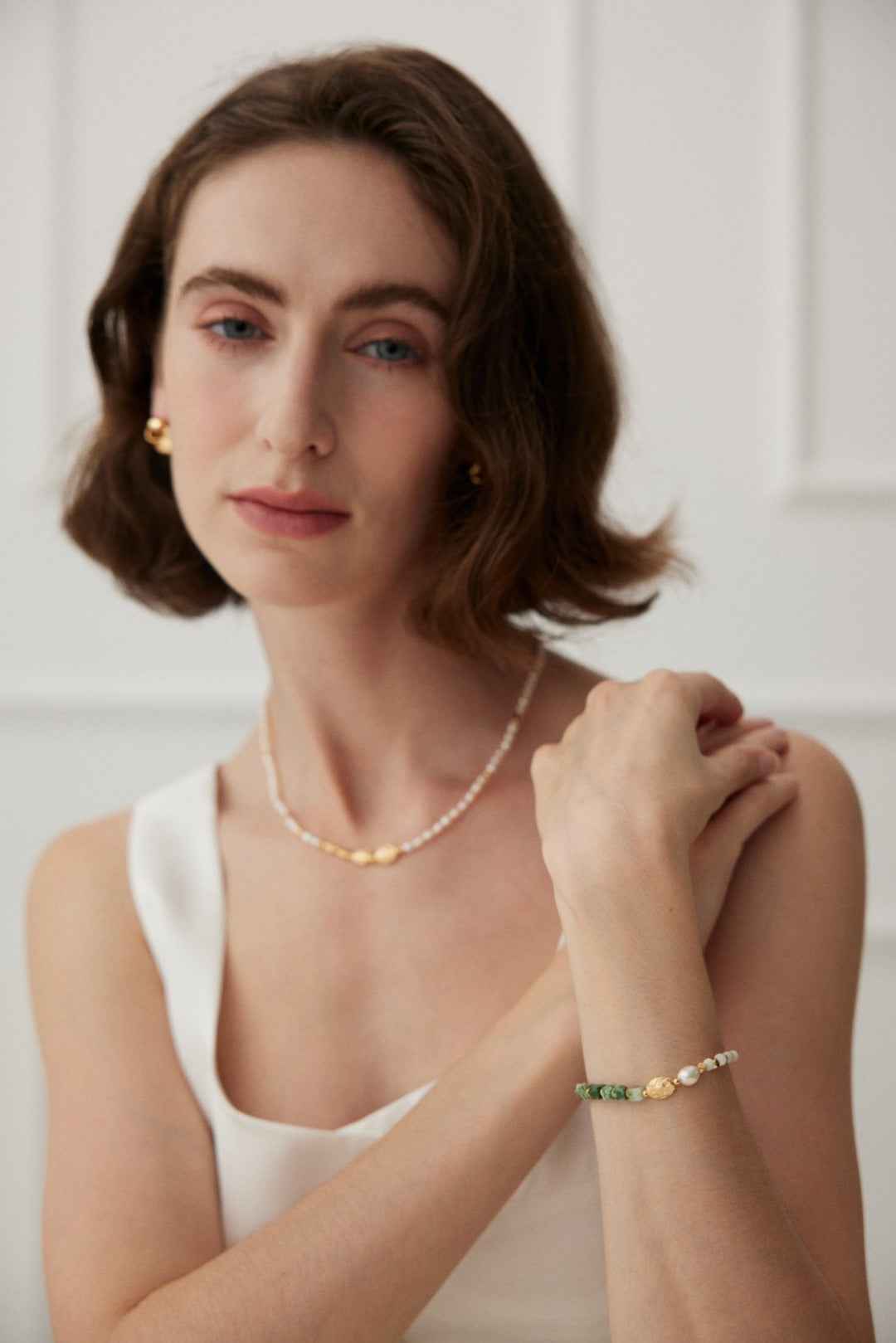 A woman with shoulder-length brown hair is wearing a sleeveless white top. She has a delicate necklace made of natural pearls and is adorned with the Elegant Vintage Gold Bracelet featuring Jade, Pearl, and Mother of Pearl in 925 Silver from Siren Mode. She looks towards the camera with a gentle expression, her arm resting on the other.