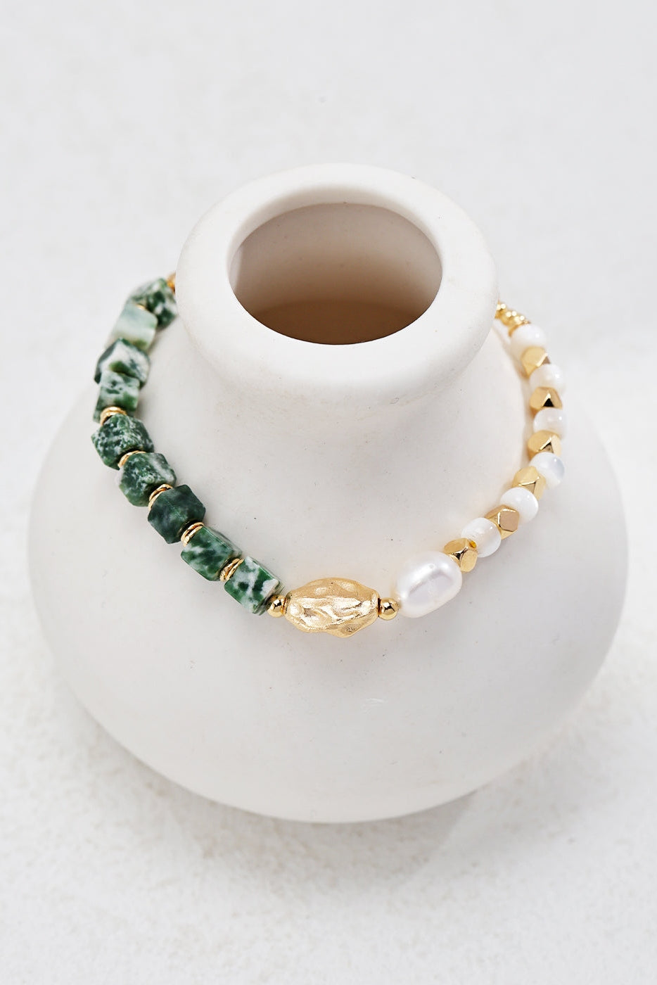 A white ceramic vase with a small opening features the Elegant Vintage Gold Bracelet from Siren Mode, draped around it. The bracelet, made of green Qinghai jade beads, gold beads, and small natural pearls set in 925 silver, creates a striking contrast against the simple and smooth surface of the vase.