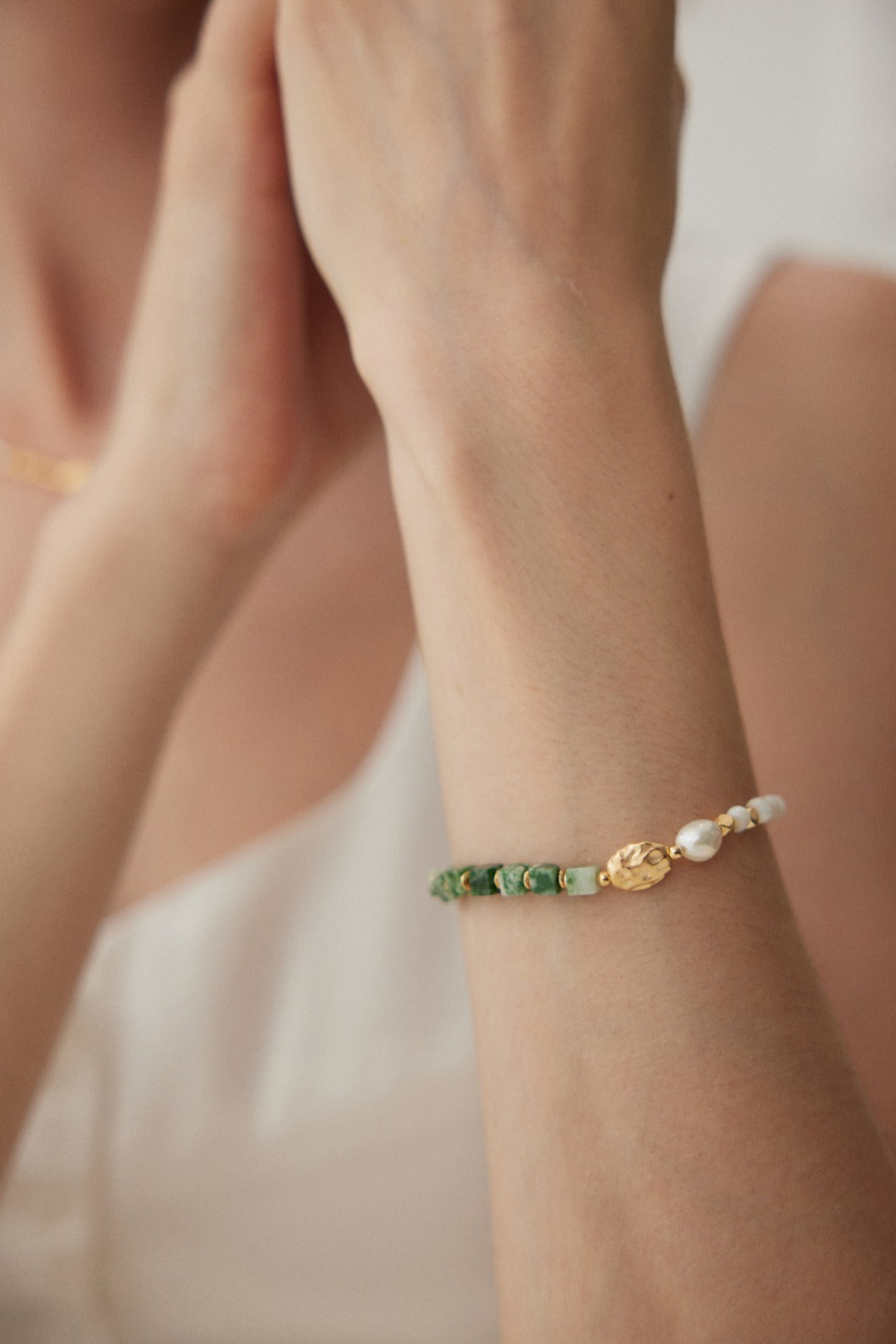 Close-up of a person wearing the Siren Mode Elegant Vintage Gold Bracelet with Jade, Pearl, and Mother of Pearl in 925 Silver on their wrist. The bracelet showcases green jade beads, natural pearls, and a small gold charm. The person is dressed in a white top, with their arm positioned near their face against a softly blurred background.