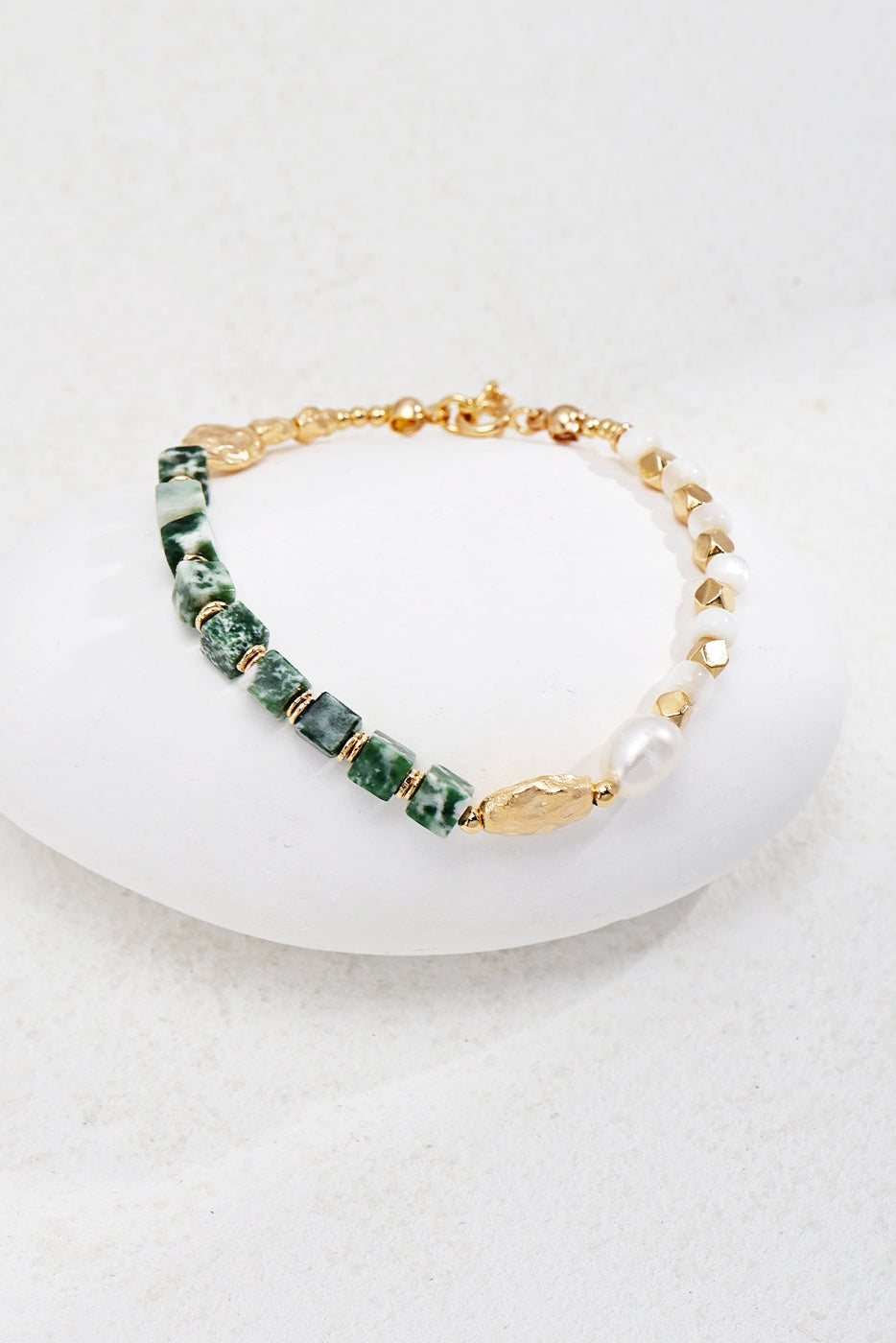 The *Elegant Vintage Gold Bracelet with Jade, Pearl, and Mother of Pearl in 925 Silver* by **Siren Mode** is beautifully showcased on a smooth white stone. This stylish accessory features alternating green Qinghai jade and gold beads, complemented by sections of varied textured beads and highlighted with a single natural pearl accent. The soft light-colored background enhances its elegant and unique design.