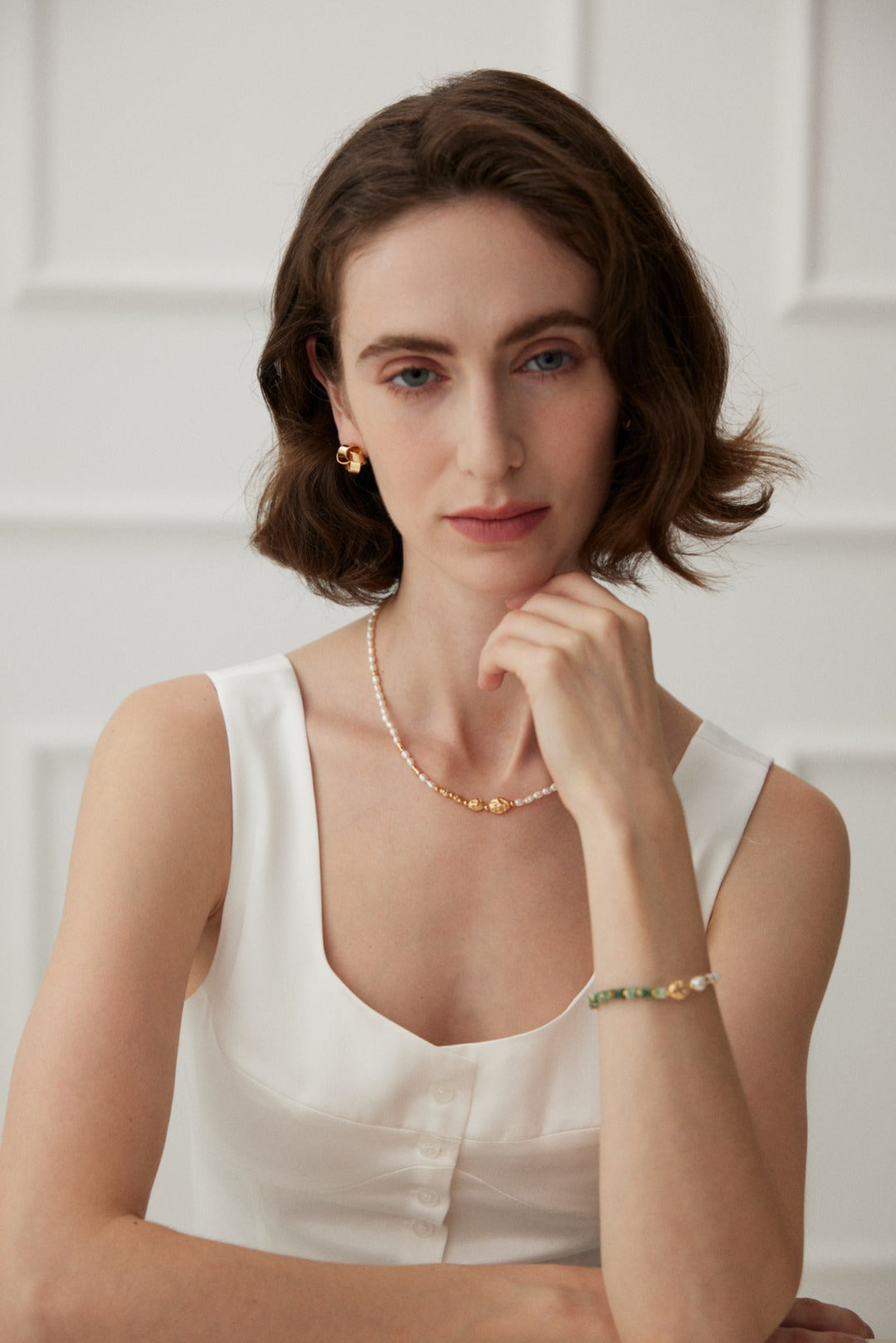 A woman with short brown hair, wearing a sleeveless white top, lightly rests her chin on her hand. Adorned with gold earrings, a delicate natural pearl necklace, and the Elegant Vintage Gold Bracelet with Jade, Pearl, and Mother of Pearl in 925 Silver by Siren Mode, she sits in front of a white paneled wall. She gazes directly at the camera with a slight smile.