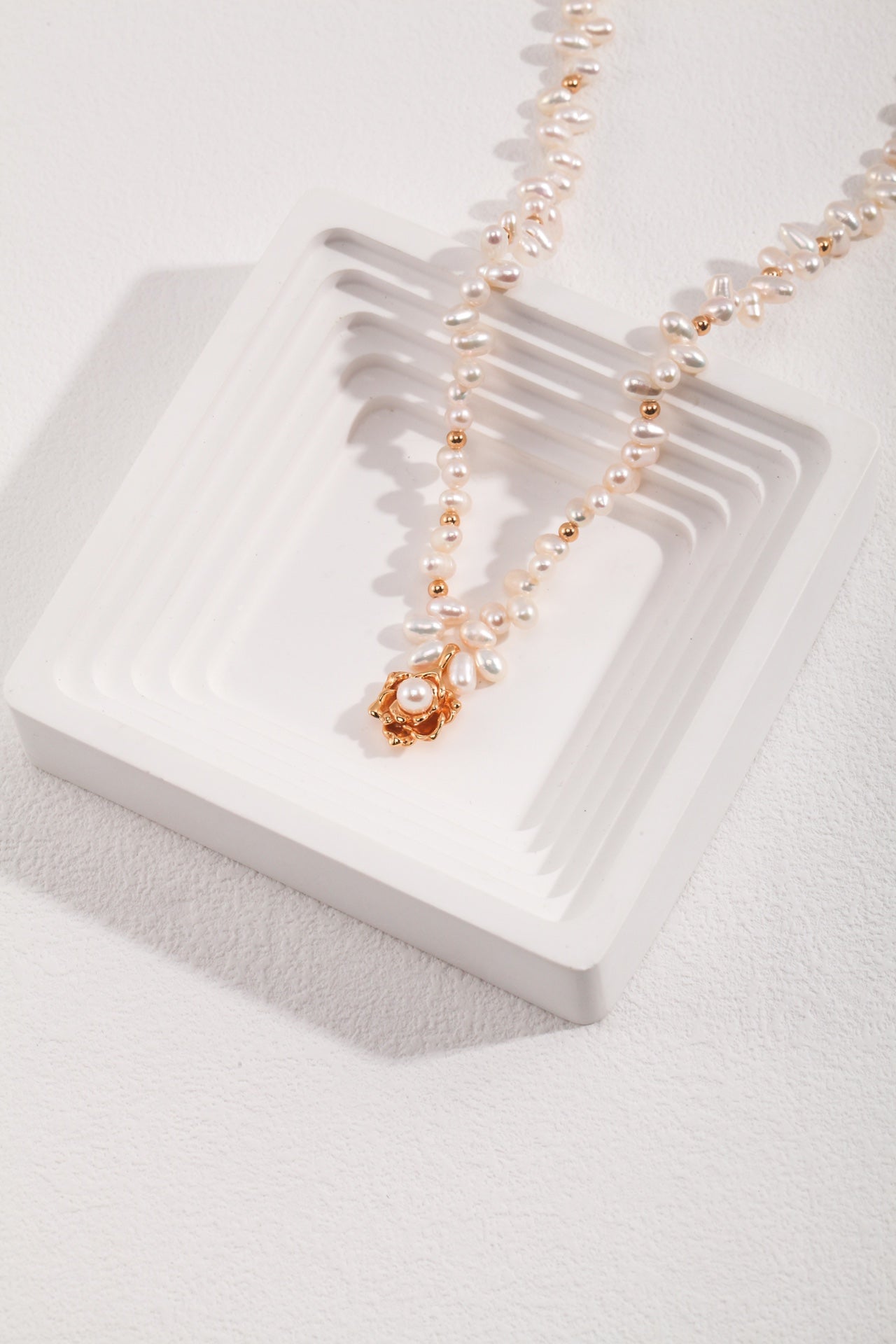 The Elegant Vintage Gold Camellia Pearl Necklace with 925 Silver Pendant by Siren Mode is displayed on a white, multi-layered square platform. Made of natural pearls complemented by golden accents, the necklace features a gold-toned pendant with a central pearl at its core. The backdrop showcases a textured white surface.