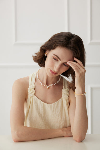 A woman with shoulder-length brown hair sits at a table wearing a light-colored sleeveless top and the Elegant Vintage Gold Camellia Pearl Necklace with a 925 Silver Pendant by Siren Mode. She rests her head on her hand with her eyes closed, appearing thoughtful or tired. The background features a white paneled wall.