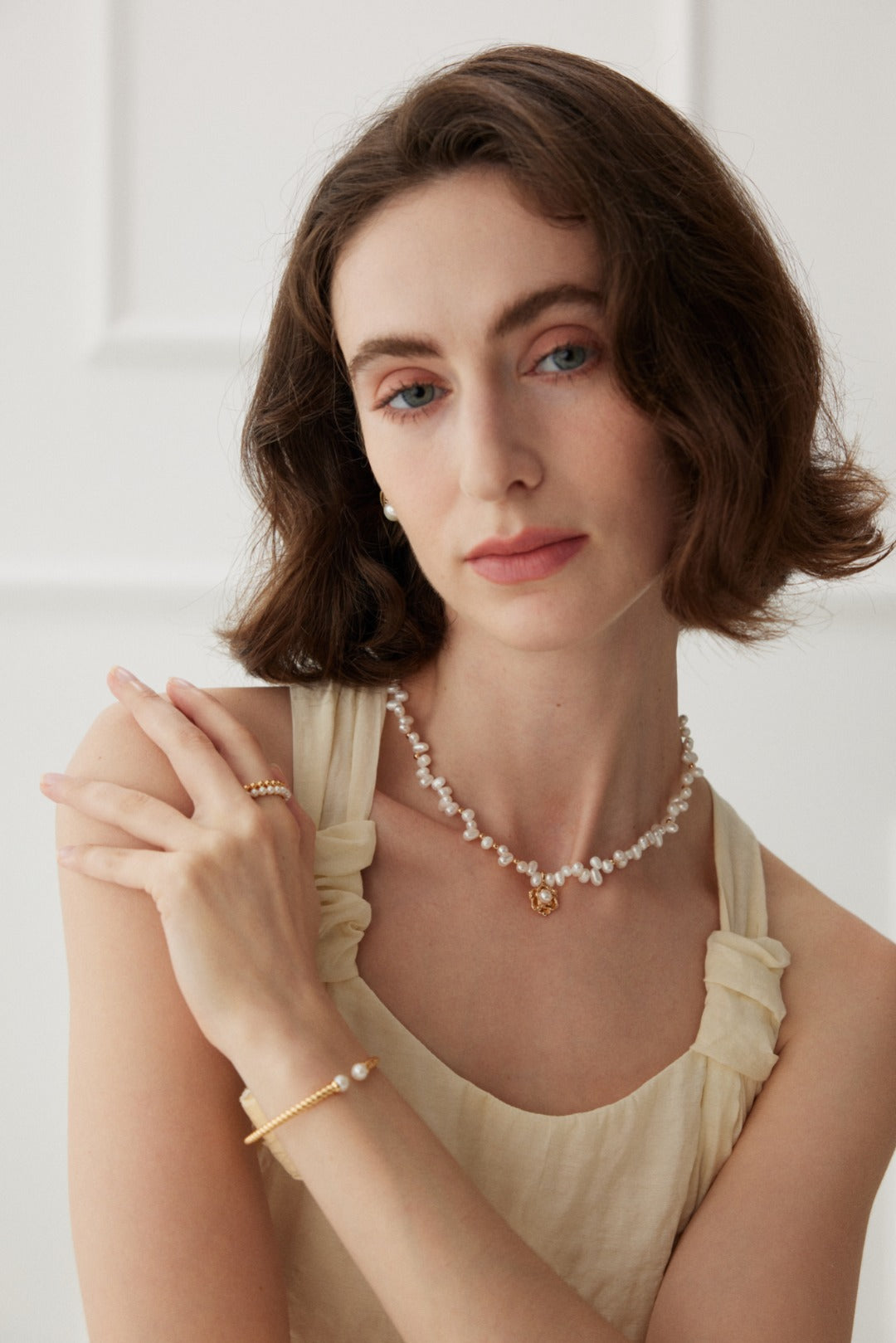 A woman with short, wavy hair is wearing a cream-colored sleeveless top. She has on the Elegant Vintage Gold Camellia Pearl Necklace with 925 Silver Pendant from Siren Mode, a pearl bracelet, and a ring on her right hand. She poses with her arms crossed gently and looks directly at the camera in a softly lit setting.