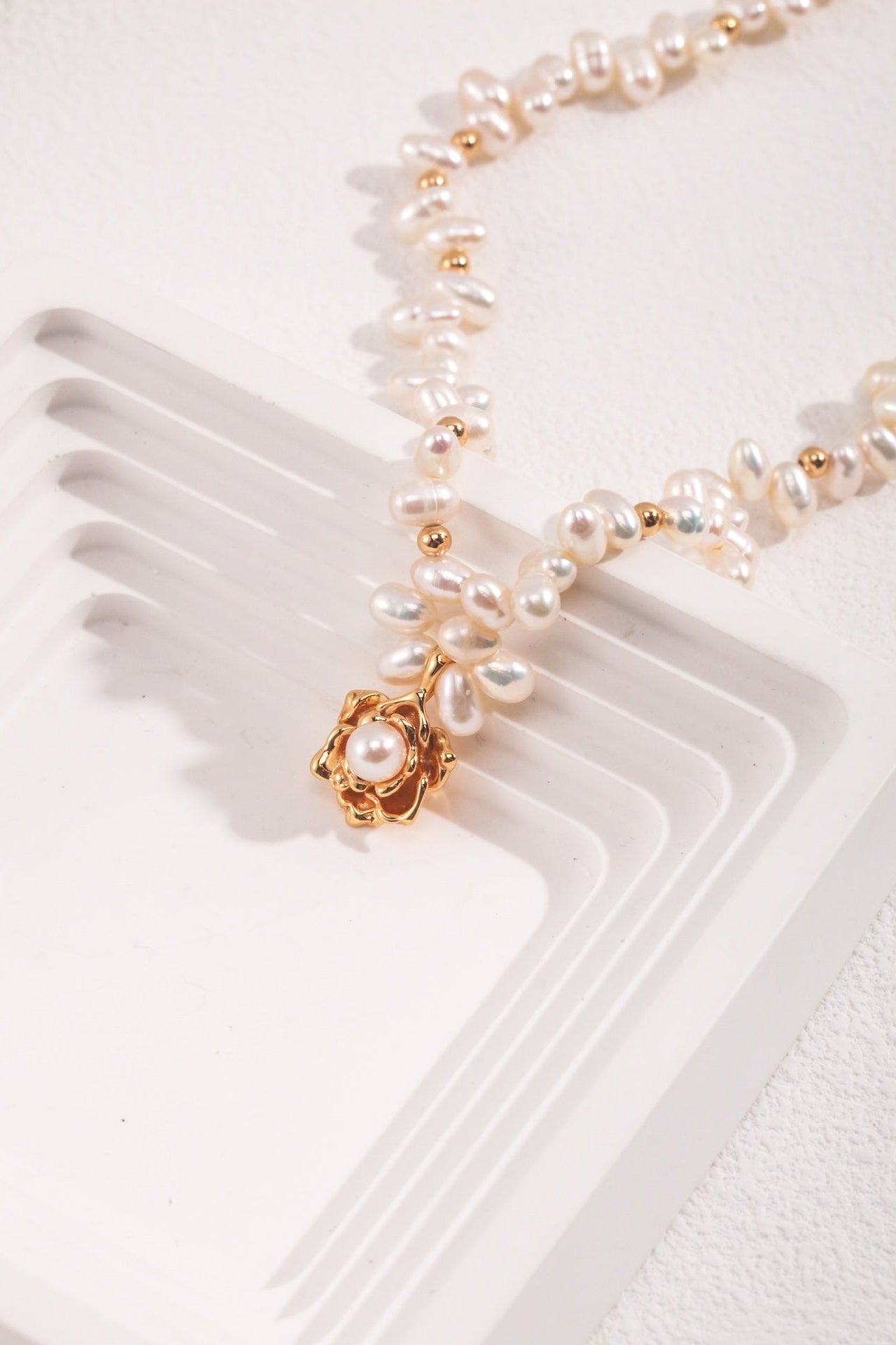 A close-up of the Elegant Vintage Gold Camellia Pearl Necklace with 925 Silver Pendant by Siren Mode, displayed on a white geometric jewelry stand with raised edges. The natural pearls are irregularly shaped, and there is a 925 silver pendant featuring a pearl at the center as the focal point of the necklace.