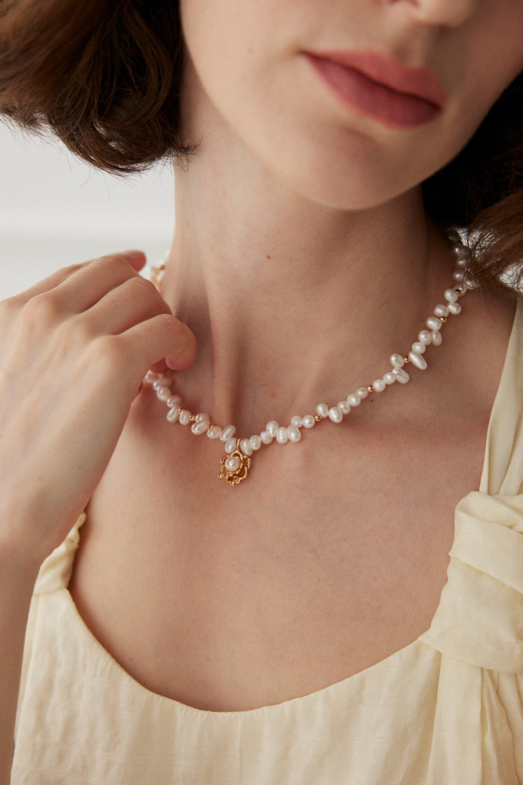 Close-up of a woman wearing the Elegant Vintage Gold Camellia Pearl Necklace with 925 Silver Pendant by Siren Mode, featuring natural pearls and a beautifully crafted pendant. Her hand gently touches the necklace as she dons a light-colored, sleeveless blouse. Her light brown hair is styled in soft waves, drawing focus to her upper body and the exquisite jewelry.