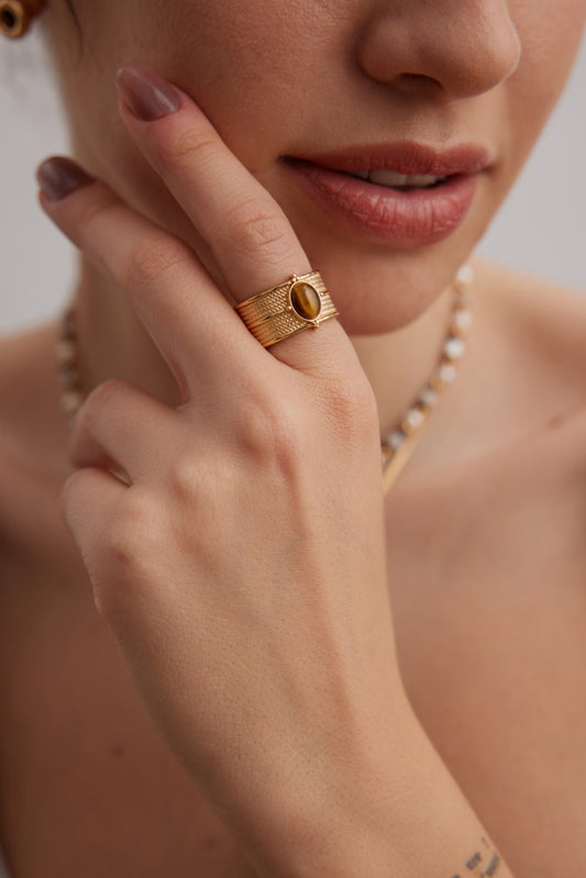 Close-up of a person's hand touching their face, adorned with Siren Mode's Elegant Vintage Gold Tiger's Eye Ring in Textured 925 Silver, featuring an oval-shaped stone. The person has light pink nails and a small tattoo on their finger. Part of a beaded necklace is visible around their neck, and their lips are slightly parted.