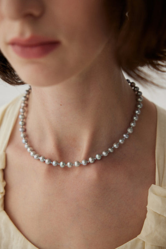 A close-up image of a woman wearing the Elegant White Gold Pearl Necklace in 925 Silver by Siren Mode. She is dressed in a light-colored top, and the focus is on her neck and the exquisite accessory. The background is softly blurred, highlighting the elegance of the pearls. Her face is partially visible, with soft, natural makeup enhancing her look.