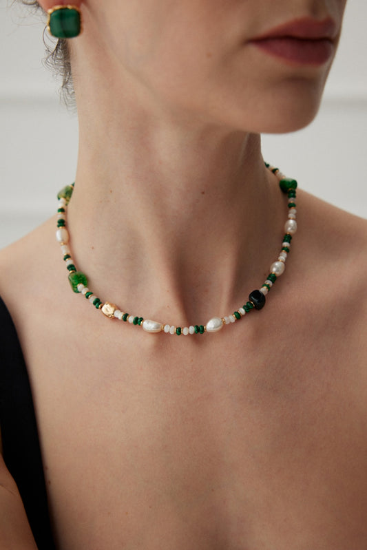 A close-up of a person wearing the Siren Mode Emerald Elegance Pearl and Agate Necklace - Vintage Gold Finish, which features green banded agate stones and irregular pearls, set in 925 silver. The individual also wears matching green earrings and is dressed in a black top. The background is softly out of focus.