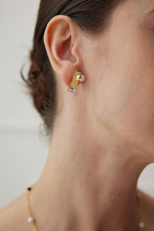 Close-up of a woman's ear adorned with Siren Mode's Geometric Pearl and Zirconia Earrings, available in Vintage Gold and White Gold. The 925 silver earring features a pearl and a small clear zirconia stone. The woman, with dark hair pulled back, has some strands visible, and she is wearing a delicate beaded necklace. The background is softly blurred.