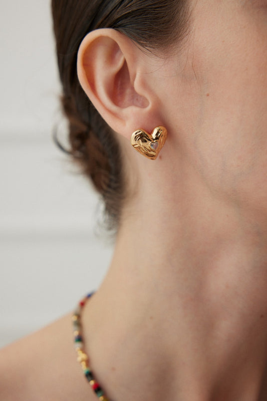 Close-up of a person wearing the Golden Love Zirconia Heart Stud Earrings by Siren Mode. Their hair is styled back, and they are also adorned with a colorful beaded necklace. The background is softly blurred, highlighting the elegance of their accessories.