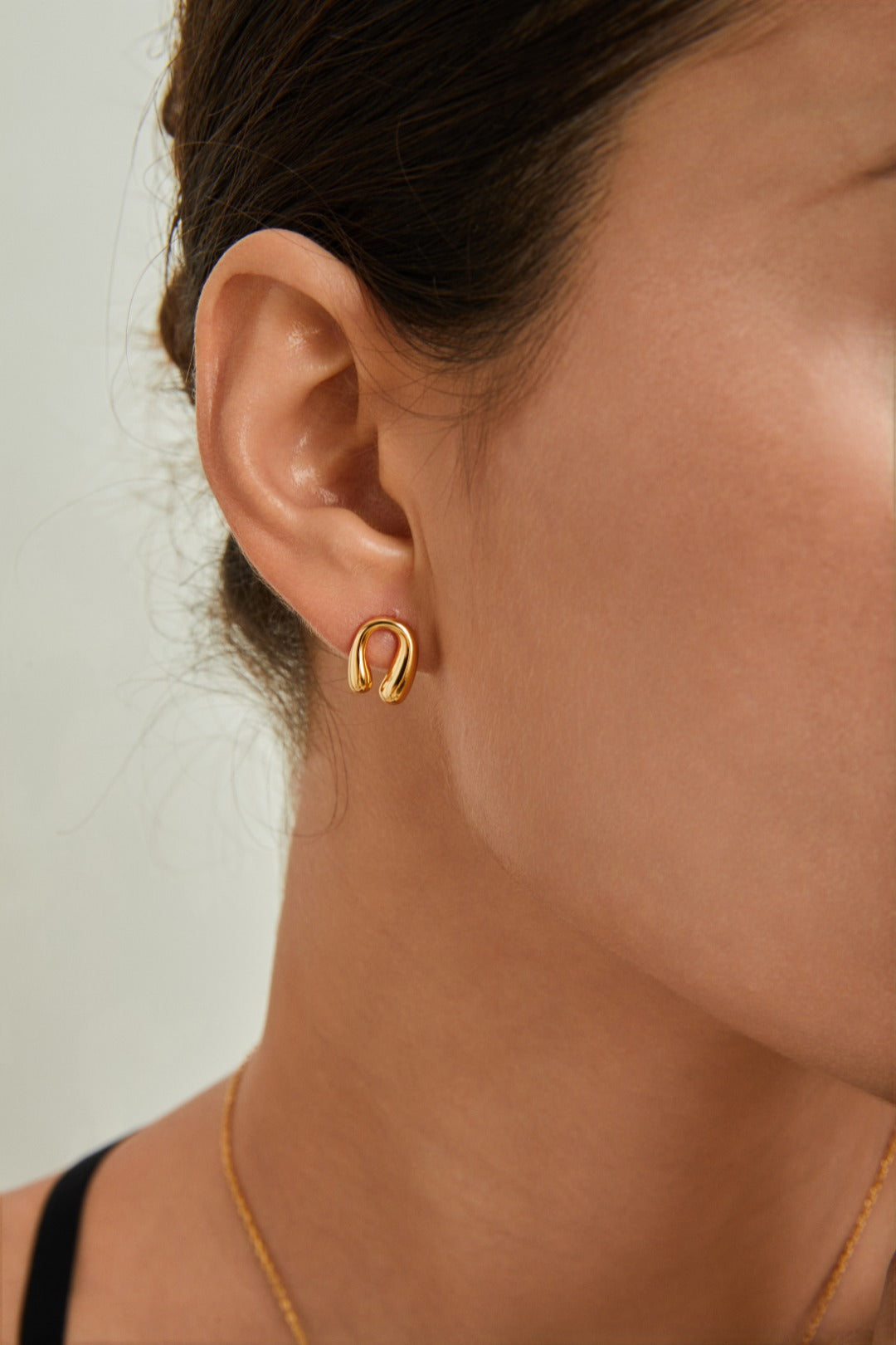 Close-up of a person wearing Siren Mode's gold-plated sterling silver Horseshoe Stud Earrings. The person's hair is pulled back, revealing the ear and part of the neck adorned with a thin necklace. The background is a muted, light gray. The overall tone of the image is neutral and elegant.