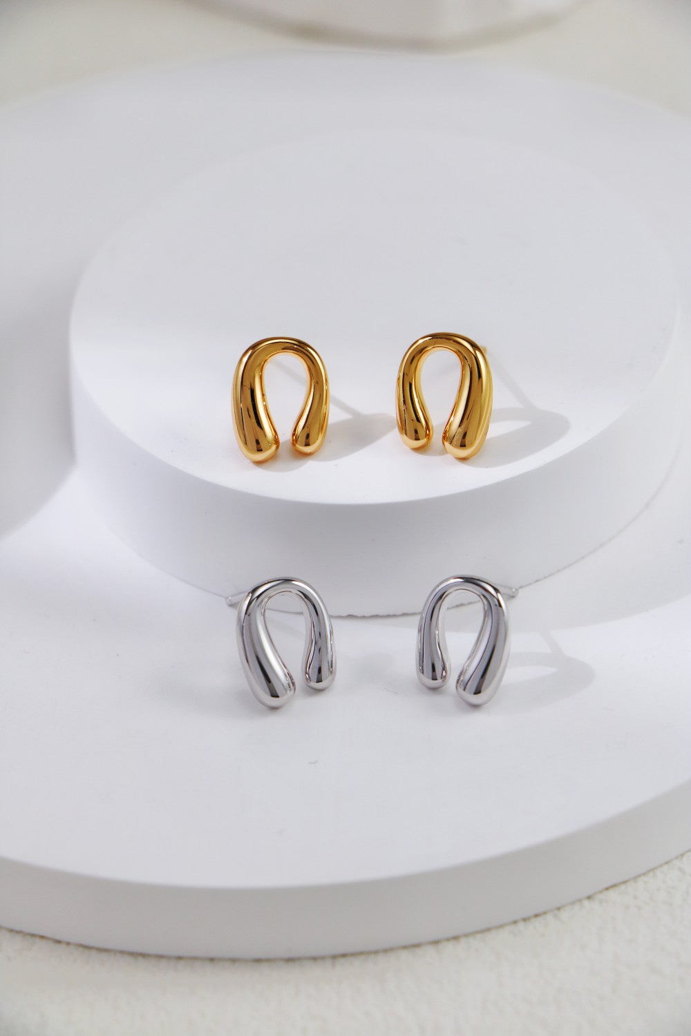 A pair of Siren Mode's Horseshoe Stud Earrings - Gold-Plated Sterling Silver and another pair in sterling silver are displayed on a round white platform. The earrings feature a modern, minimalist design with an open loop shape. The image exudes a clean and elegant aesthetic, capturing the understated charm for which horseshoe stud earrings are known.