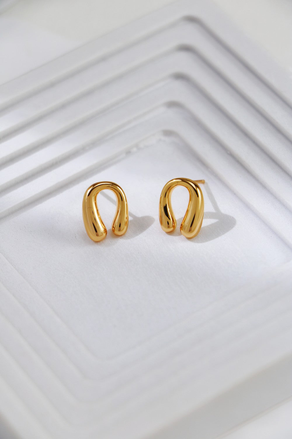 A pair of Siren Mode Horseshoe Stud Earrings in gold-plated sterling silver is placed on a white textured surface with layered, square-shaped ridges. The metallic shine of the earrings contrasts with the matte, bright background.