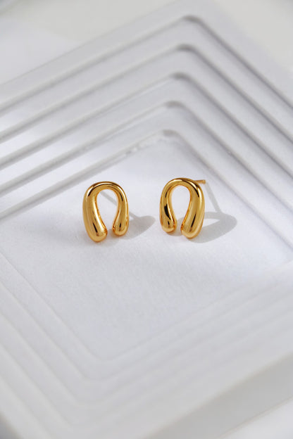 A pair of Siren Mode Horseshoe Stud Earrings in gold-plated sterling silver is placed on a white textured surface with layered, square-shaped ridges. The metallic shine of the earrings contrasts with the matte, bright background.