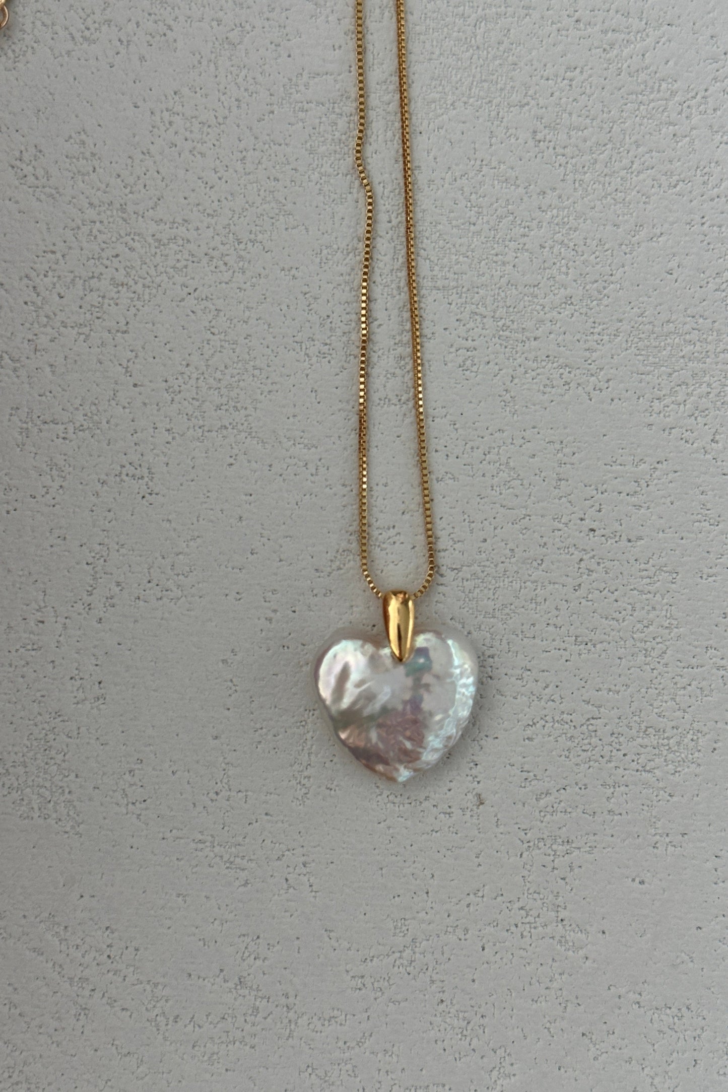 The Large Heart Pendant Necklace by Siren Mode features a sophisticated 18K gold-plated, sterling silver chain with a heart-shaped pendant adorned in a pearlescent, iridescent finish and secured with a 14K gold clasp. The Baroque Pearl Heart is beautifully showcased against a textured, light gray background.