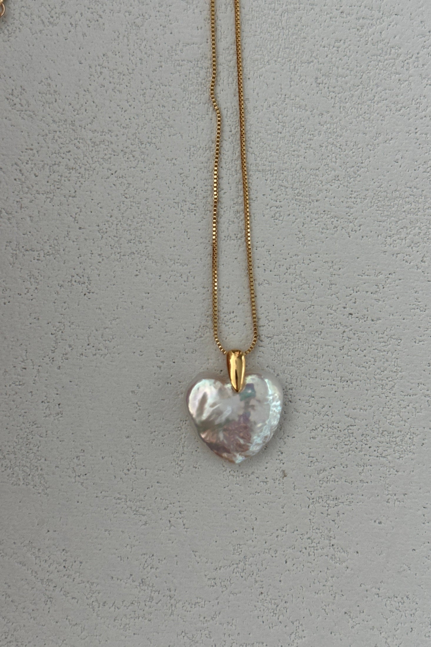 The Large Heart Pendant Necklace by Siren Mode features a sophisticated 18K gold-plated, sterling silver chain with a heart-shaped pendant adorned in a pearlescent, iridescent finish and secured with a 14K gold clasp. The Baroque Pearl Heart is beautifully showcased against a textured, light gray background.