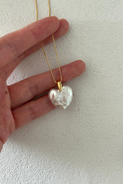 A hand holds the Siren Mode Large Heart Pendant Necklace, an 18K gold-plated sterling silver chain with a Baroque Pearl Heart pendant, its iridescent, pearly material shimmering. The background is a textured, off-white surface.