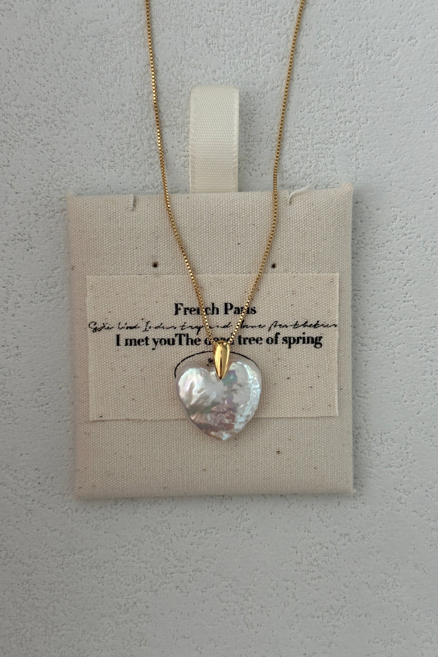 The Siren Mode Large Heart Pendant Necklace, featuring an 18K gold-plated sterling silver baroque pearl heart pendant with a 14K gold clasp, is elegantly displayed on a beige fabric pouch. The pouch features faint black text reading "French Paris. I met you The breeze of spring" against a textured light gray wall background.