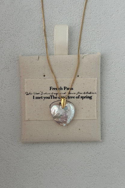 The Siren Mode Large Heart Pendant Necklace, featuring an 18K gold-plated sterling silver baroque pearl heart pendant with a 14K gold clasp, is elegantly displayed on a beige fabric pouch. The pouch features faint black text reading "French Paris. I met you The breeze of spring" against a textured light gray wall background.